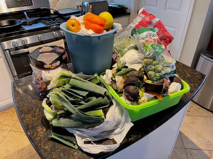 A kitchen island covered with bags and pails of food scraps including cucumbers, avocado skins, bell peppers, leak leaves, etc.